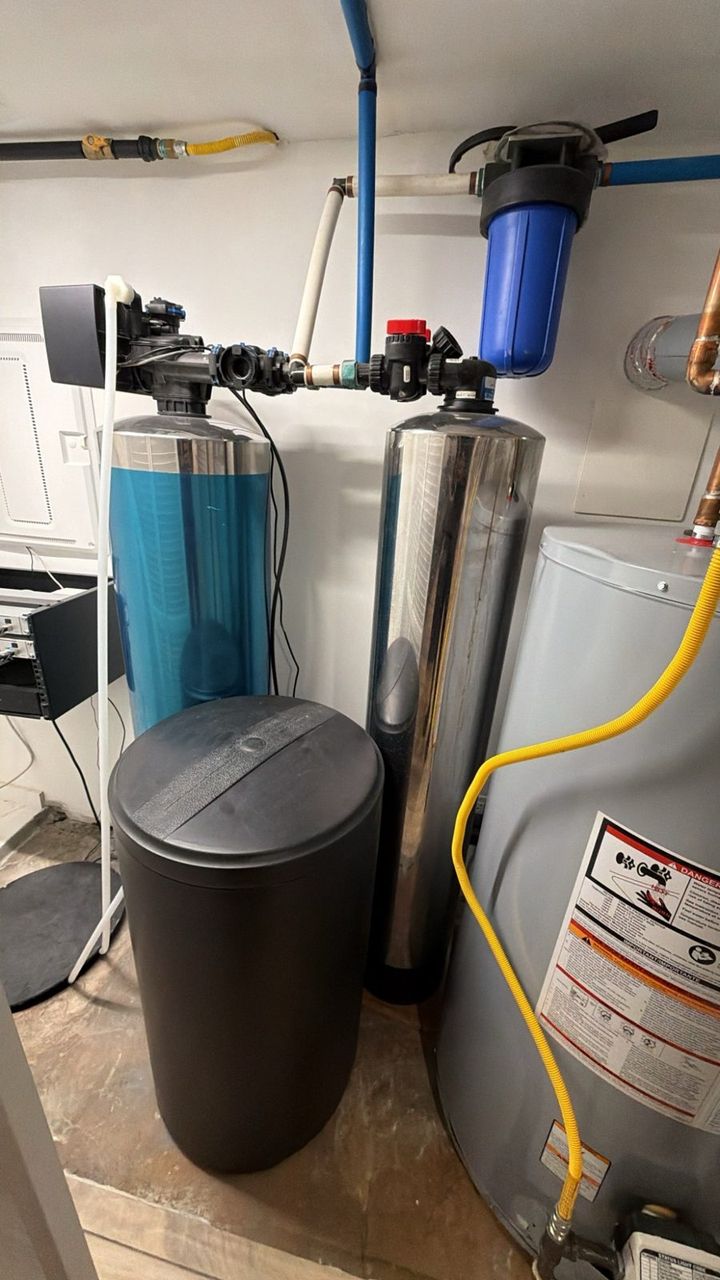 A blue-tinted water softener on the left with a black salt tank in front of it, and a tall stainless carbon filter tank on the right, with a blue sediment filter on the pipe above and a water heater beside them.